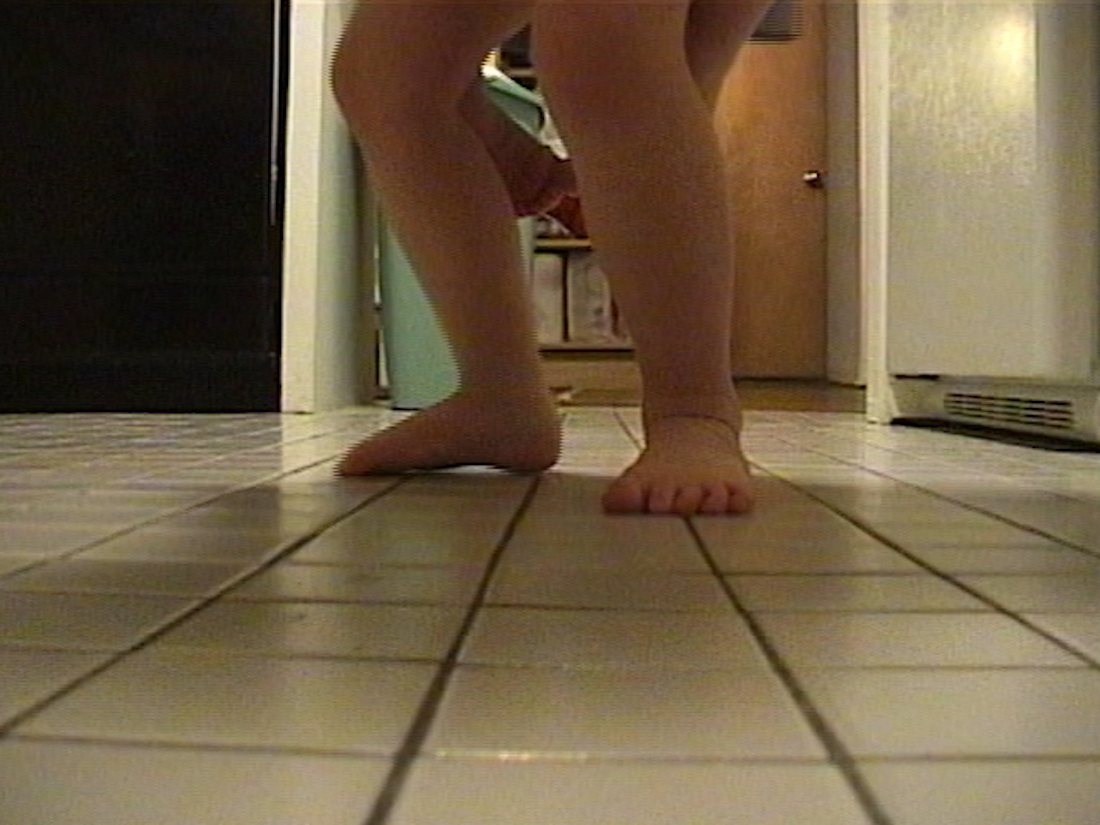 A barefoot baby seen from the knees down, standing on a tile floor