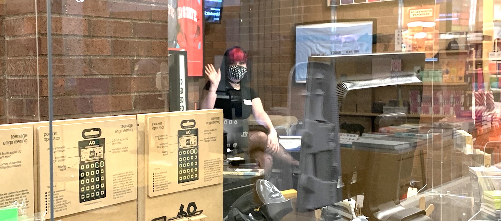 A young white woman in black with bright pink bangs and a studded face mask waves to the camera from behind a store counter displaying various items