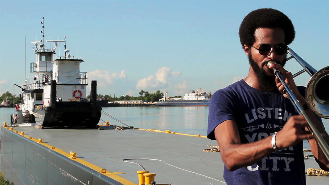 A man of color in a graphic t-shirt and sunglasses plays a trombone on the far right side of the frame, standing on a pier with a tugboat and body of water in the background