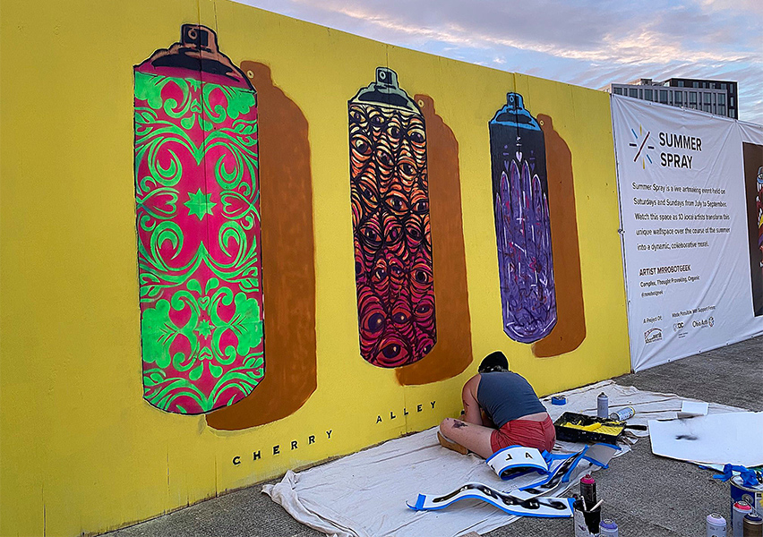 Artist Katie Golonka is seen from the side as she completes a mural of three pattern-embellished spray paint cans against a solid field of bright yellow