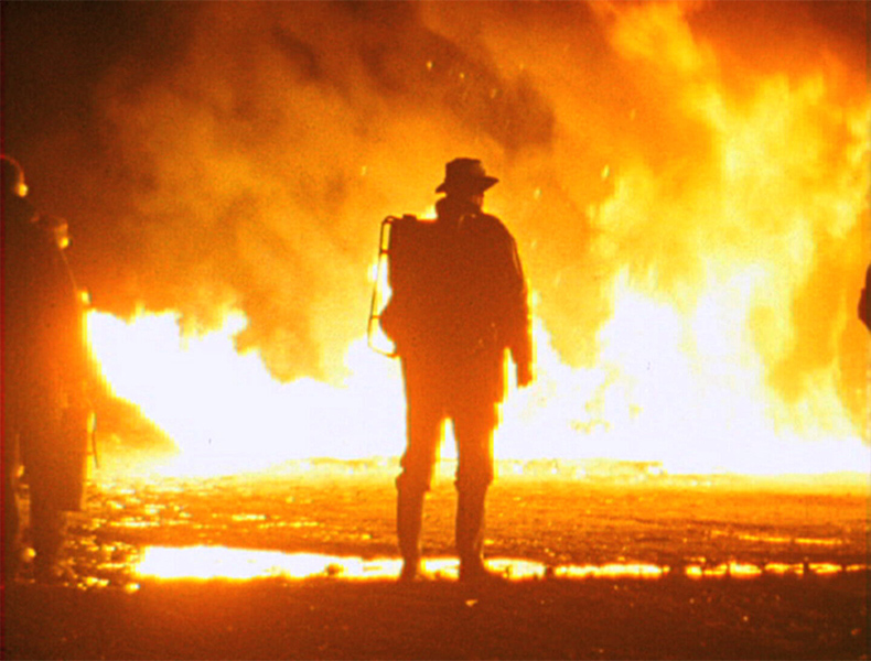 The back of a firefighter seen in silhouette in a night scene with a wall of flames in the background