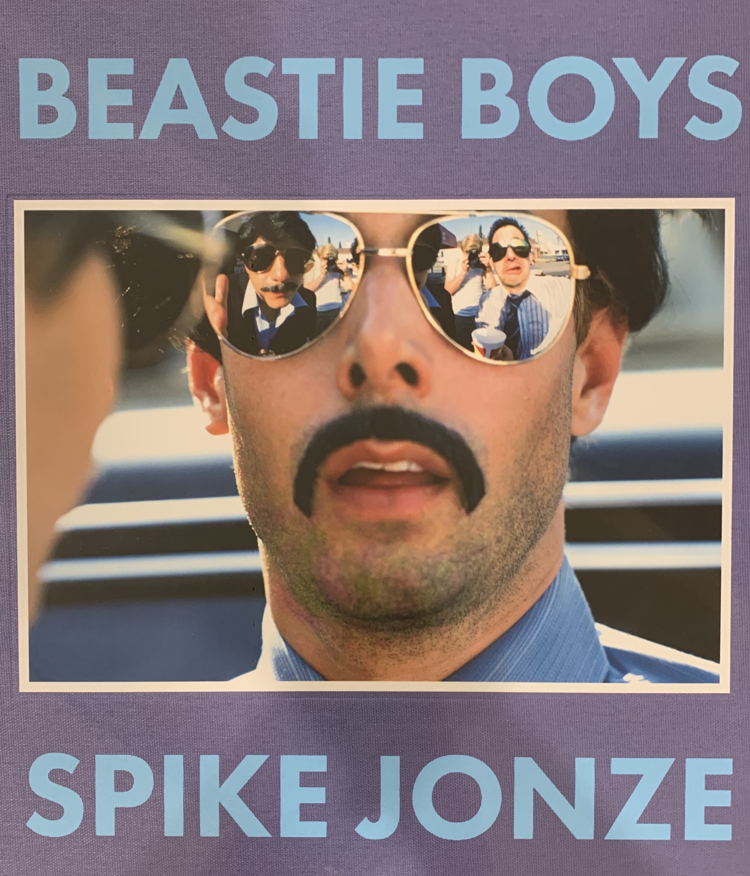 A member of the Beastie Boys dressed as a police officer with mirrored sunglasses and a fake mustache is pictured on the cover of a book about the band by Spike Jonze. Title and author names are in light blue on purple background at top and bottom of photo