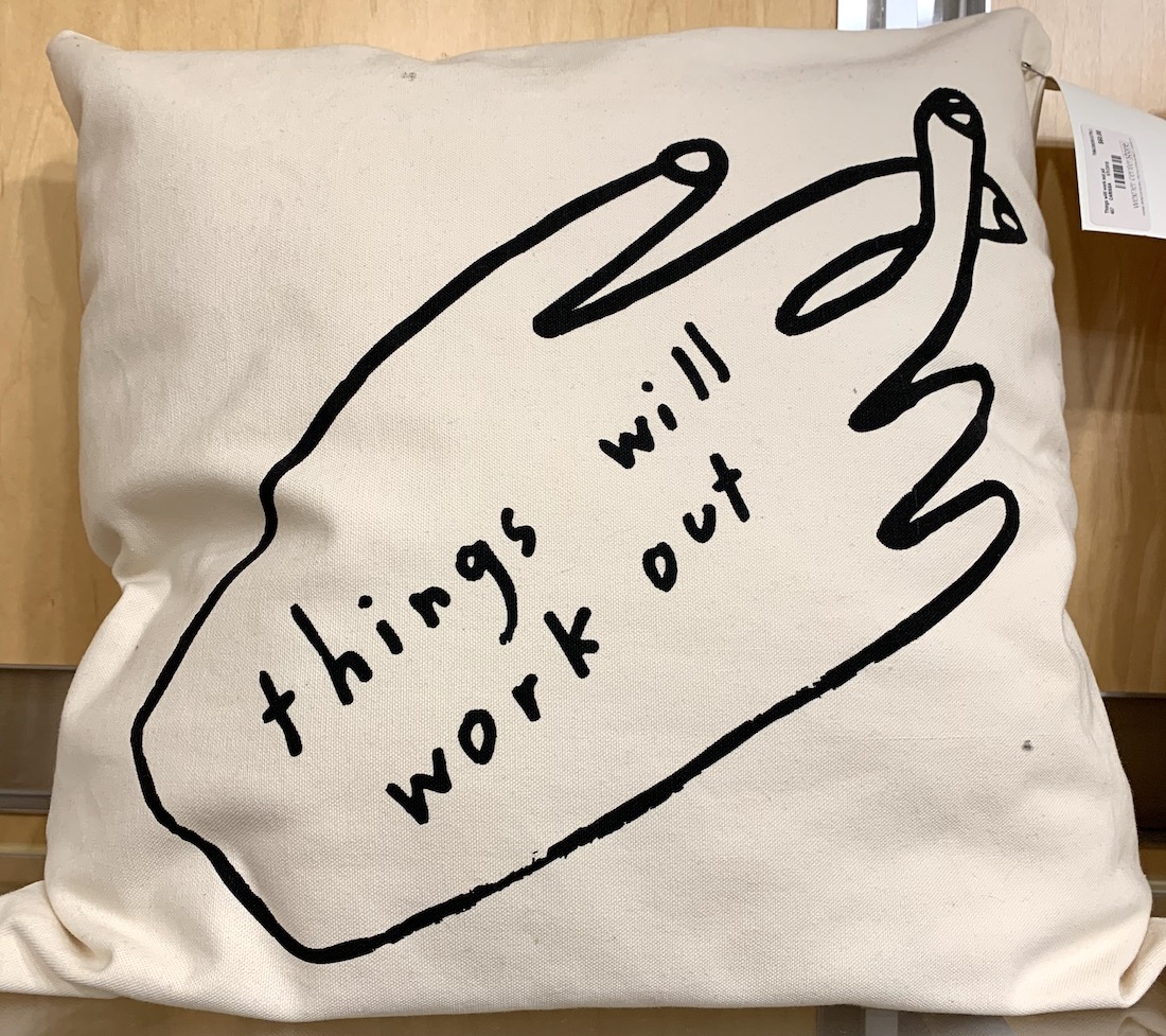 An off white pillow with a drawing of a right hand with crossed fingers and "things will work out" written on the hand's back