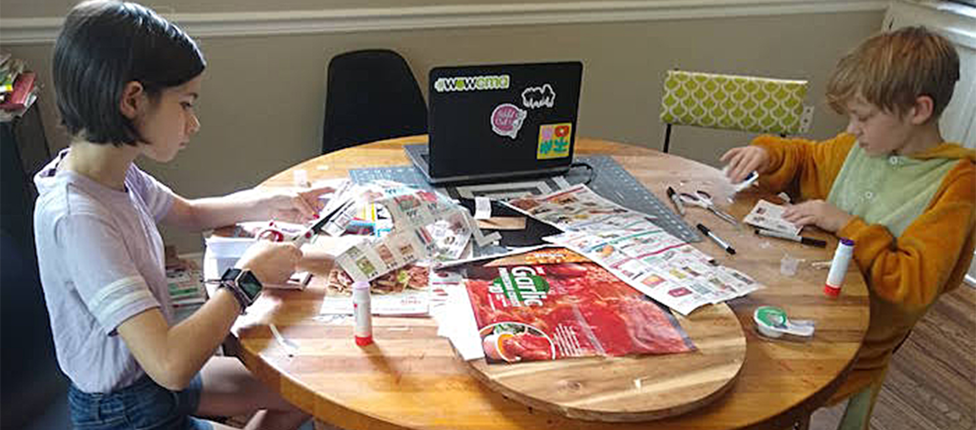 A young girl and boy sit and work at a table covered in printed circulars, flattened packaging, and art supplies