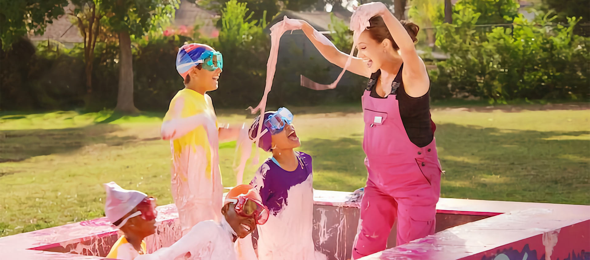 A woman in pink overalls plays with four children in a small outdoor pool filled with pink slime
