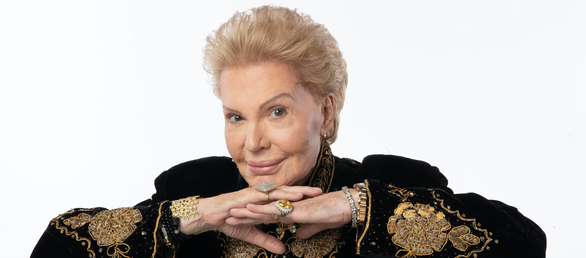 Astrologer Walter Mercado folds his hands below his chin, wearing a gold embroidered black jacket against a white background