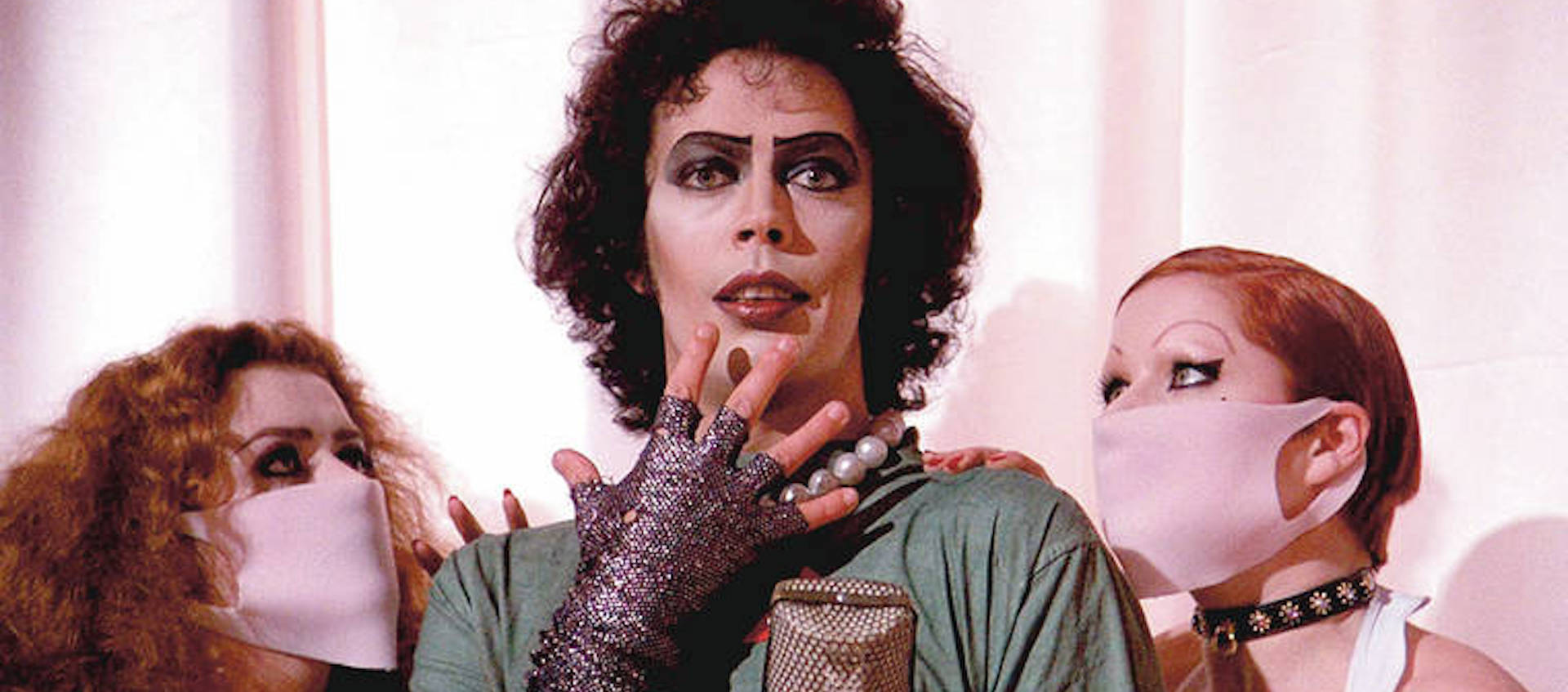 Tim Curry's transvestite Dr. Frank N. Furter stands in front of a white curtain, flanked by nurses in white face masks, in a scene from the film The Rocky Horror Picture Show