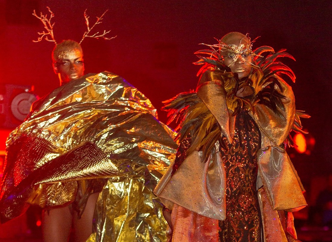 Two Black women with short cropped hair wear elaborate metallic costumes, makeup and hair accessories on a red-lit stage as part of a runway show during Highball Halloween