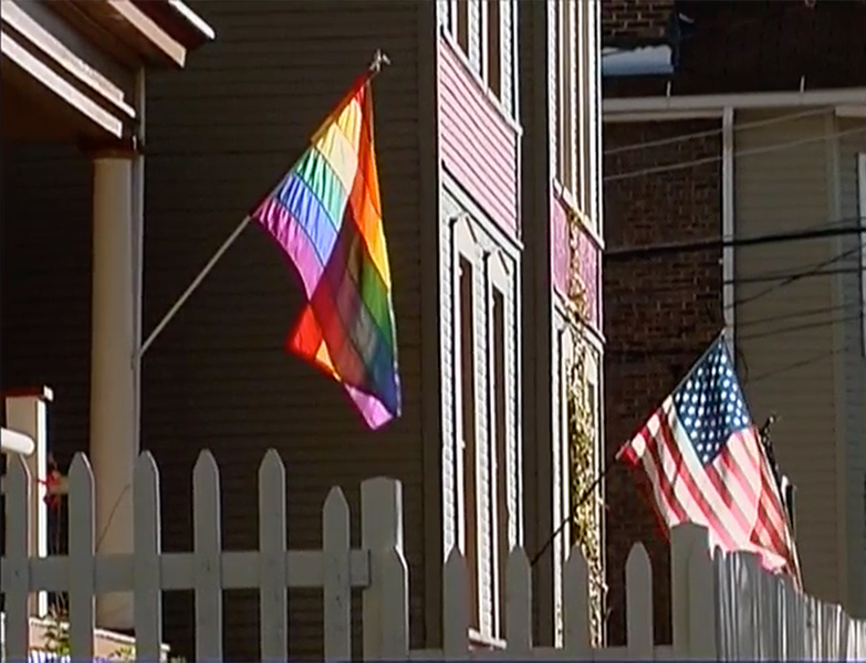 Pride flag hanging on the porch of a home. Next to this house is a home with an American flag hanging in front.