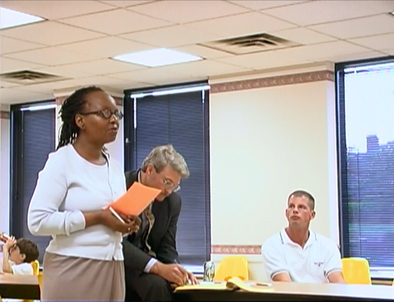 In the left of the image, a woman stands addressing someone in the room. She is holding an orange piece of paper. To her left are two men, one standing and one seated looking at her.