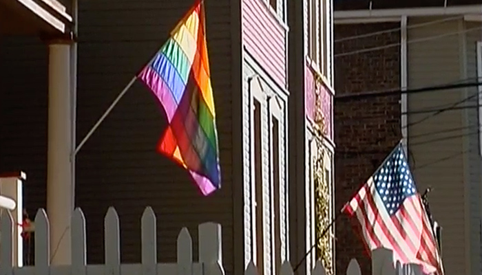 Pride flag hanging on the porch of a home. Next to this house is a home with an American flag hanging in front.