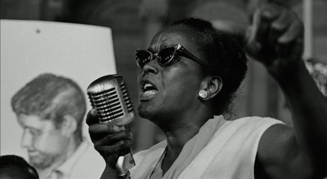 Image of Ella Baker wearing sunglasses and standing at a microphone speaking passionately