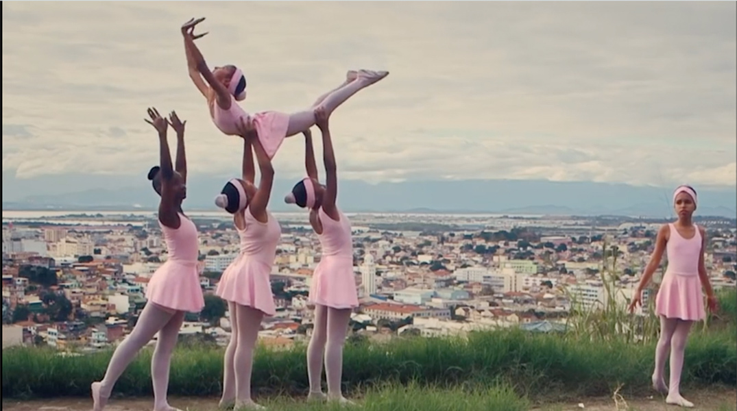 On top of a hill overlooking a cityscape, three young women in pink tutus lift another young woman in a pink tutu above their heads. To the far right another young woman in a pink tutu stands looking ahead.
