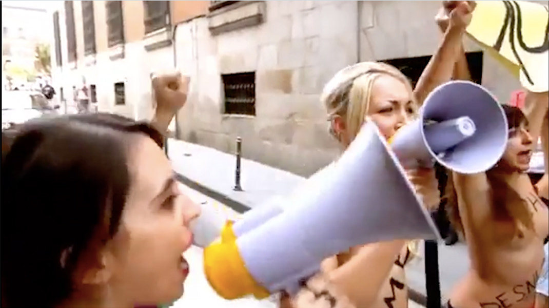 A group of three shirtless women stand together speaking through megaphones