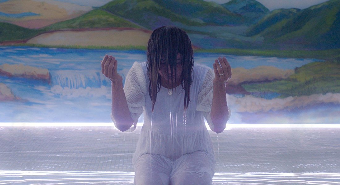 A woman dressed all in white kneels in a pool of water. Her arms are aloft and she is soaked in water.