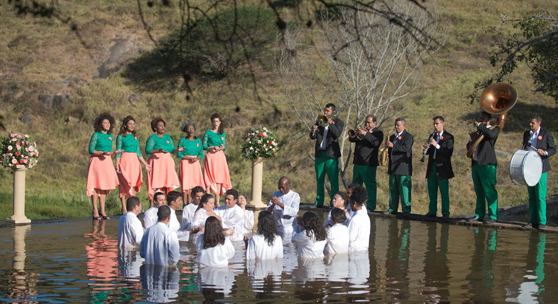 A group of people stand up to their waists in water in a baptismal ceremony. On the shore stand two separate groups, on the right are musicians playing instruments, on the left are four people in matching green shirts and pink skirts.