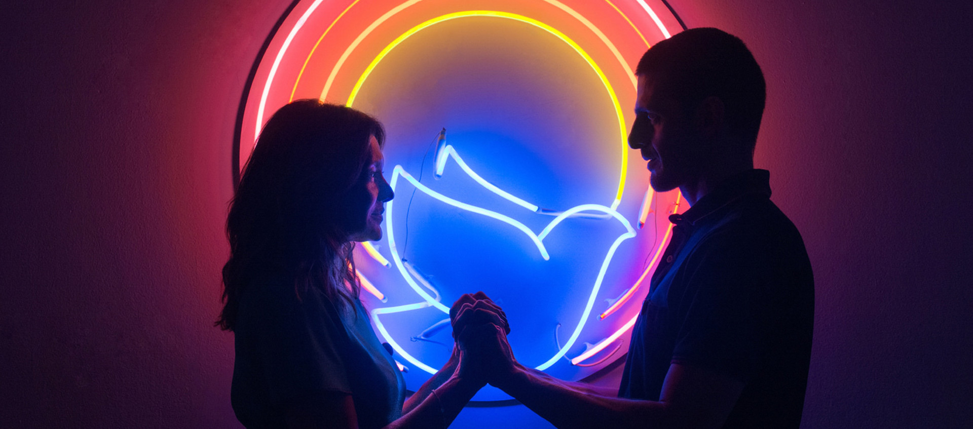 Two silhouettes, a woman on the left and a man on the right, holding hands, standing in front of a neon circle that features a blue neon dove in the center