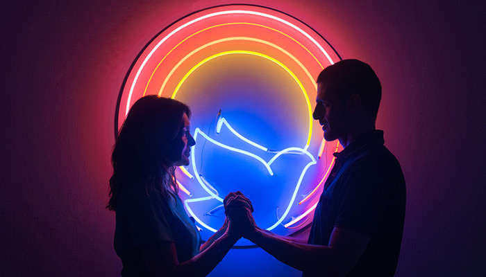Two silhouettes, a woman on the left and a man on the right, holding hands, standing in front of a neon circle that features a blue neon dove in the center