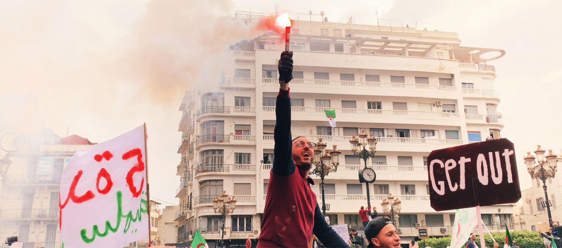 Man at protest standing in front of a building while he holds up a flare