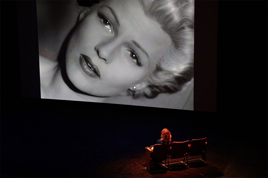 Nina Menkes sits on stage and watches a black and white film depicting a woman's face that fills the frame. 