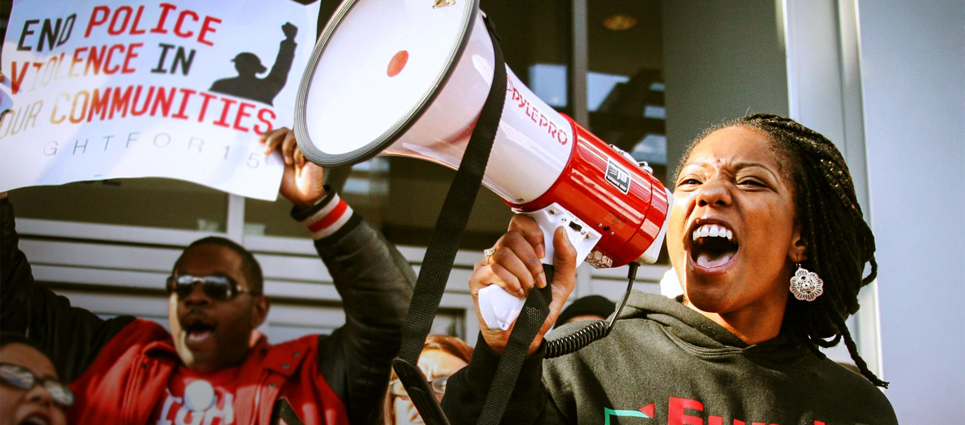 A woman shouts passionately into a megaphone while a man behind her holds up a sign supporting the end of police violence.