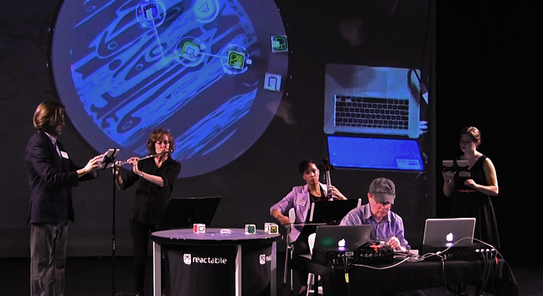 Five musicians onstage, from left a man uses an iPad, a woman plays flute, a seated woman plays cello, in foreground a man sits at a laptop, and on far right a woman uses an iPad