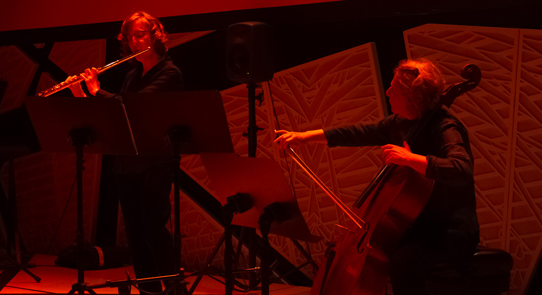 Two musicians bathed in red light, on the left one plays a flute on the right one is seated playing the cello