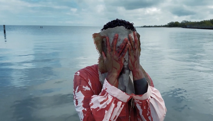 A black trans woman standing in Lake Pontchartain in a white tunic covered in blood uses a towel to wipe blood from their face