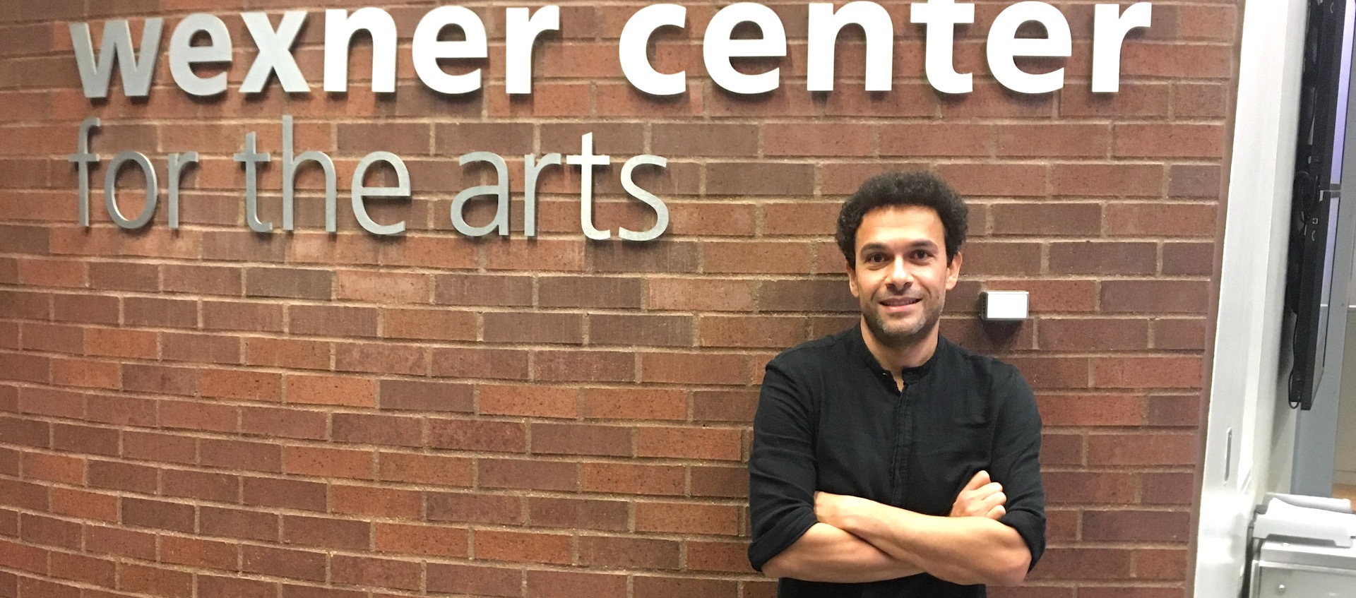 Filmmaker Tamer El Said stands against a brick wall with the Wexner Center for the Arts logo in brushed metal lettering above him