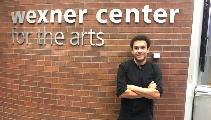 Filmmaker Tamer El Said stands against a brick wall with the Wexner Center for the Arts logo in brushed metal lettering above him
