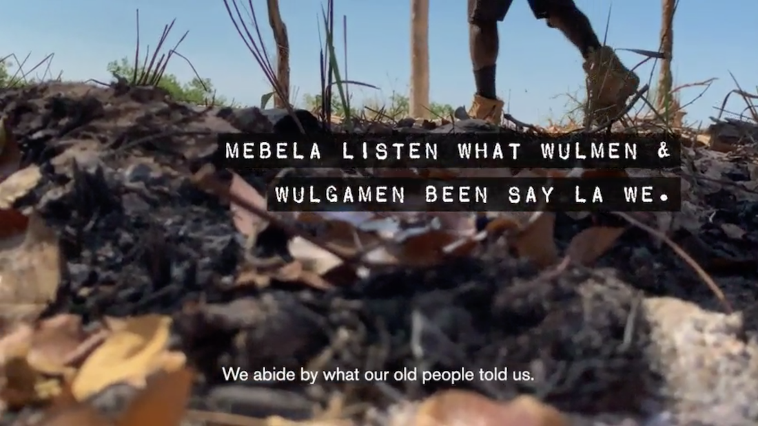 A man's boot-covered feet seen walking across a wooded area with a graphic overlaid with white text on black bars: "Mebela listen what wulmen and wulgamen been say la we," translated below the text in English: "We abide by what our old people told us."