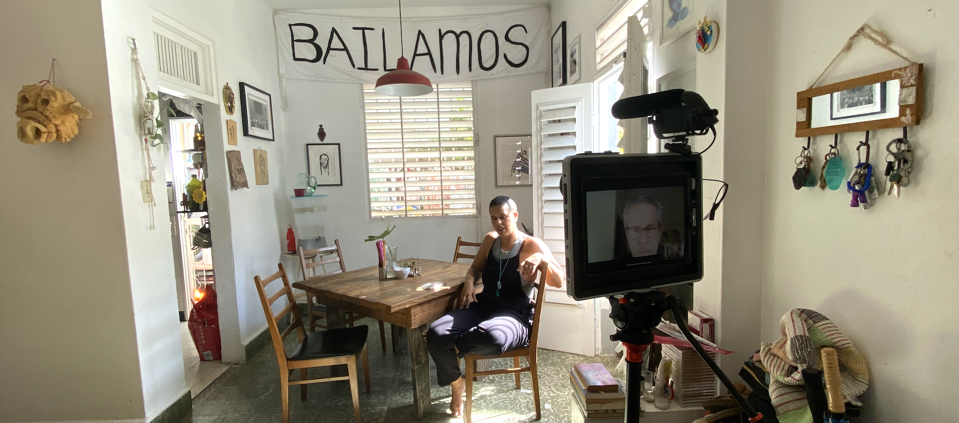 Choreographer Awilda Rodriguez Lora sits at a small, square dining table in her living and performance space La Rosario in Puerto Rico. A video camera recording her can be seen to the right in the forefront of the frame.