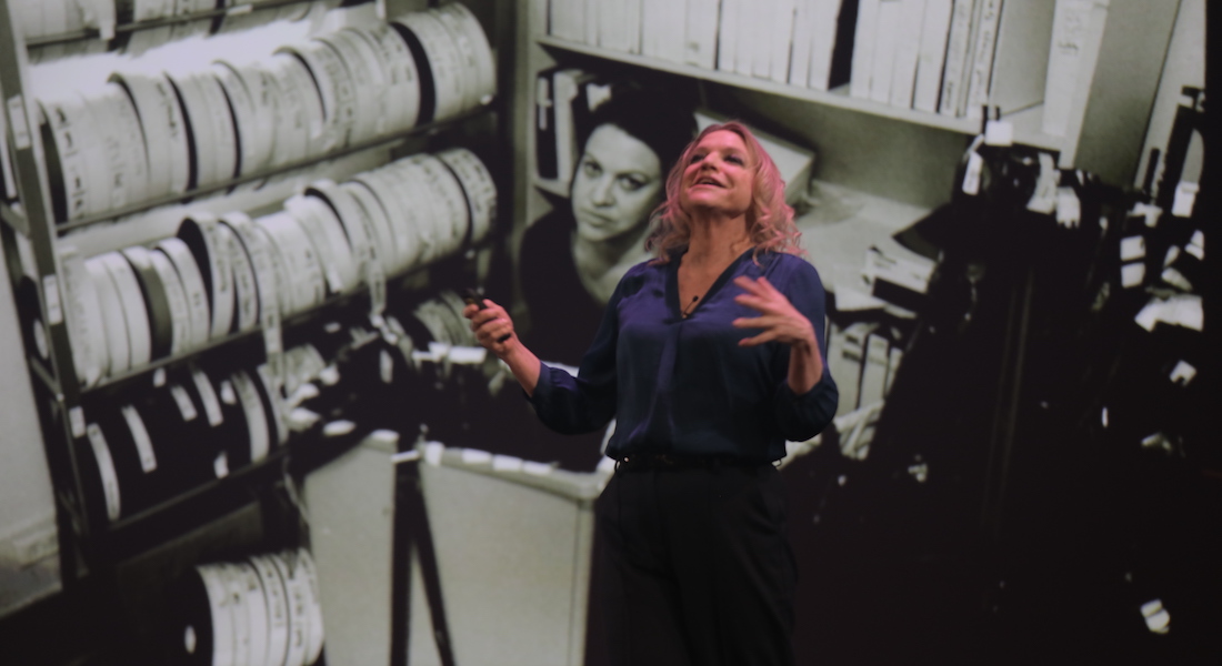 Filmmaker Nina Menkes stands in front of a projection screen filled with a vintage black and white image of a woman film editor at work, surrounded by reels of film