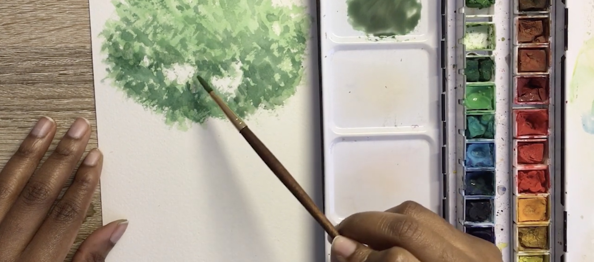 On a wood work surface with an open container of watercolor paints, a black woman's hands apply green paint to white paper, creating the look of a tree top