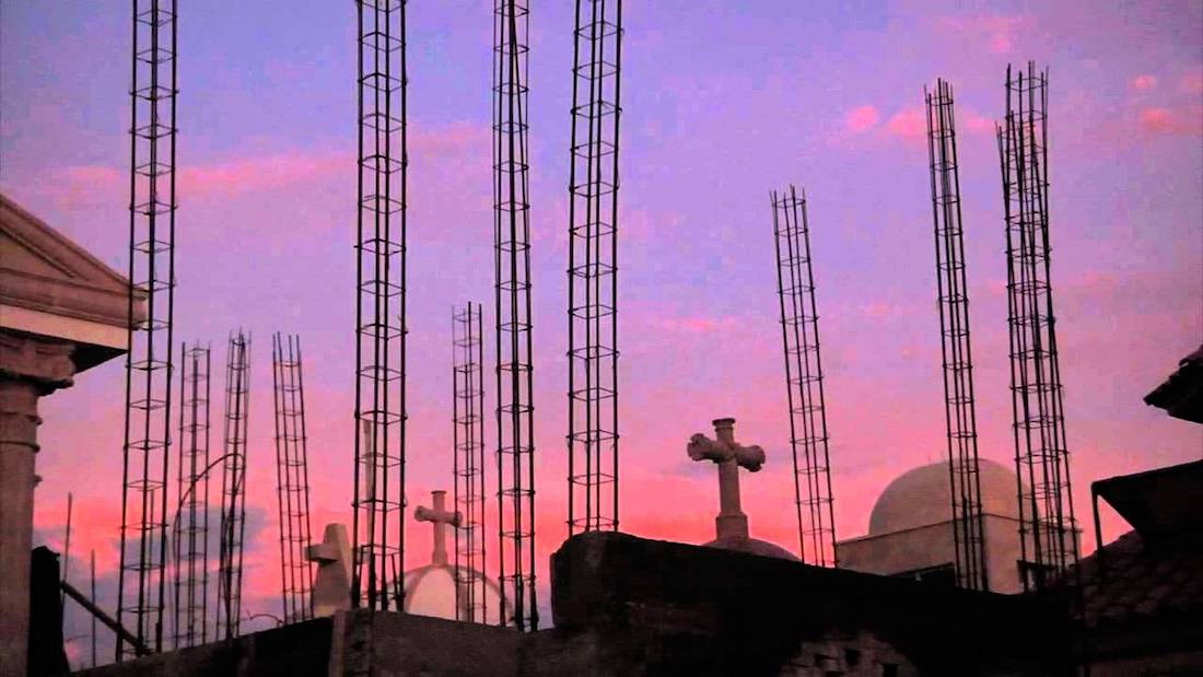 A rooftop scene of crosses and rebar towers against a purple and pink sunset