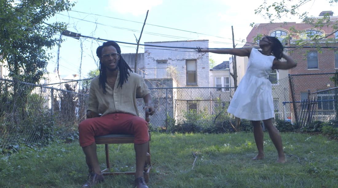 On the left, in an outdoor, grassy setting with buildings in the background, a Black man with long braids sits in a chair singing as a Black woman with long braids in a white dress dances on the right side