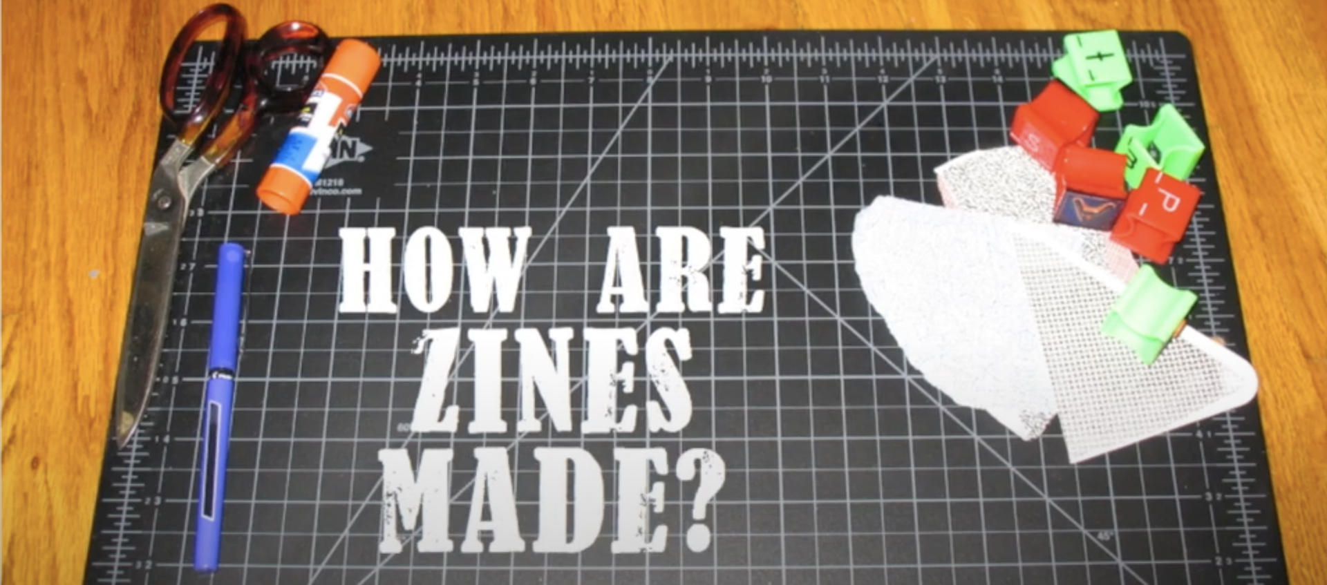 Image of a grid-covered cutting board on a wood table, covered in drawing and cutting tools and marked by the text graphic "How are zines made?"