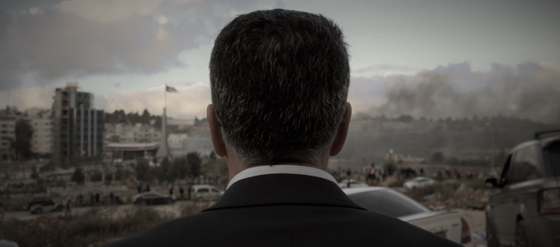 A man with dark hair in a suit is seen from behind in the foreground; the man is looking out at a city skyline under a gray, cloudy sky