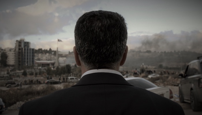 A man with dark hair in a suit is seen from behind in the foreground; the man is looking out at a city skyline under a gray, cloudy sky