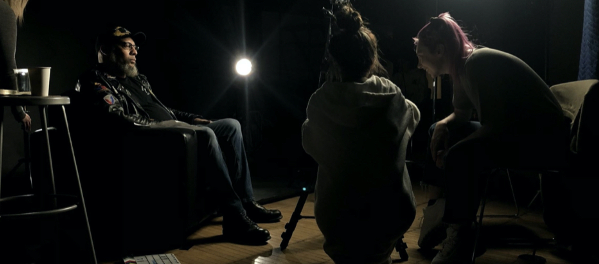 Artist Leni Anderson sits to the left as he's recorded on video during an interview with two young filmmakers, seen on the right from the side in a dimly lit shooting space