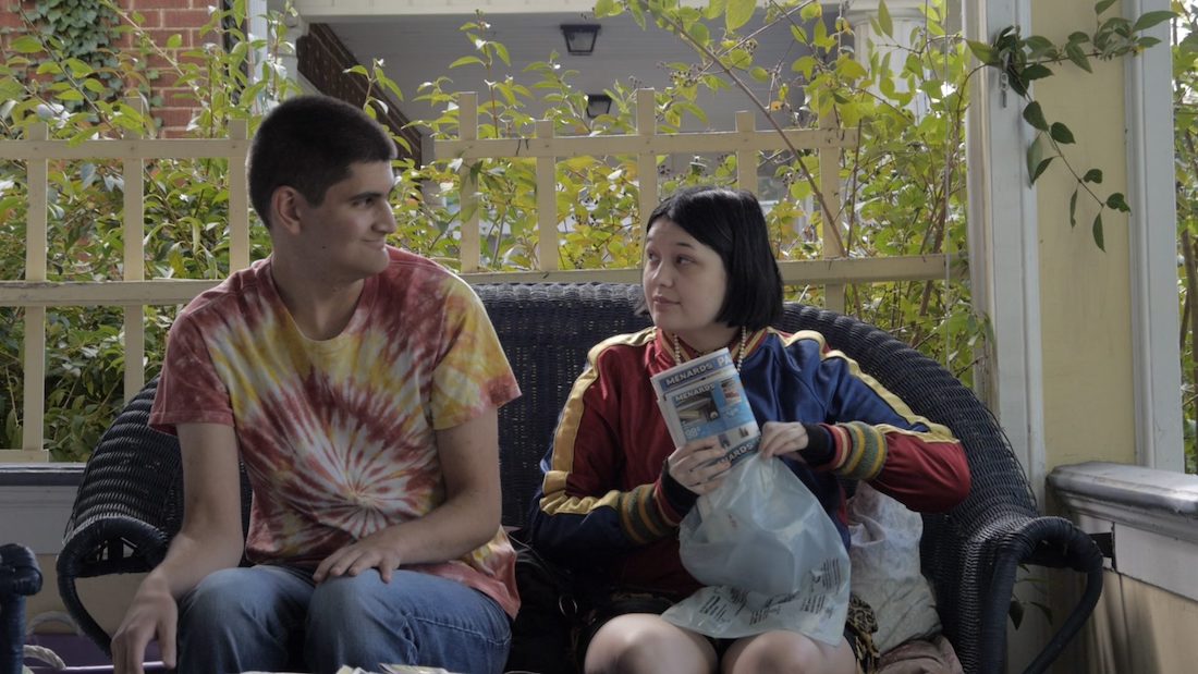 A still from the film Those Who Spring of Me. A young white man and young white woman sit beside each other on a rattan couch on a porch with leafy trees in the background. He has short, dark hair. He is wearing a tie dye T-shirt and is looking at the woman while leaning slightly away from her. She has short, straight hair and is wearing a red, yellow and blue satin jacket. She appears to be saying something to the man.