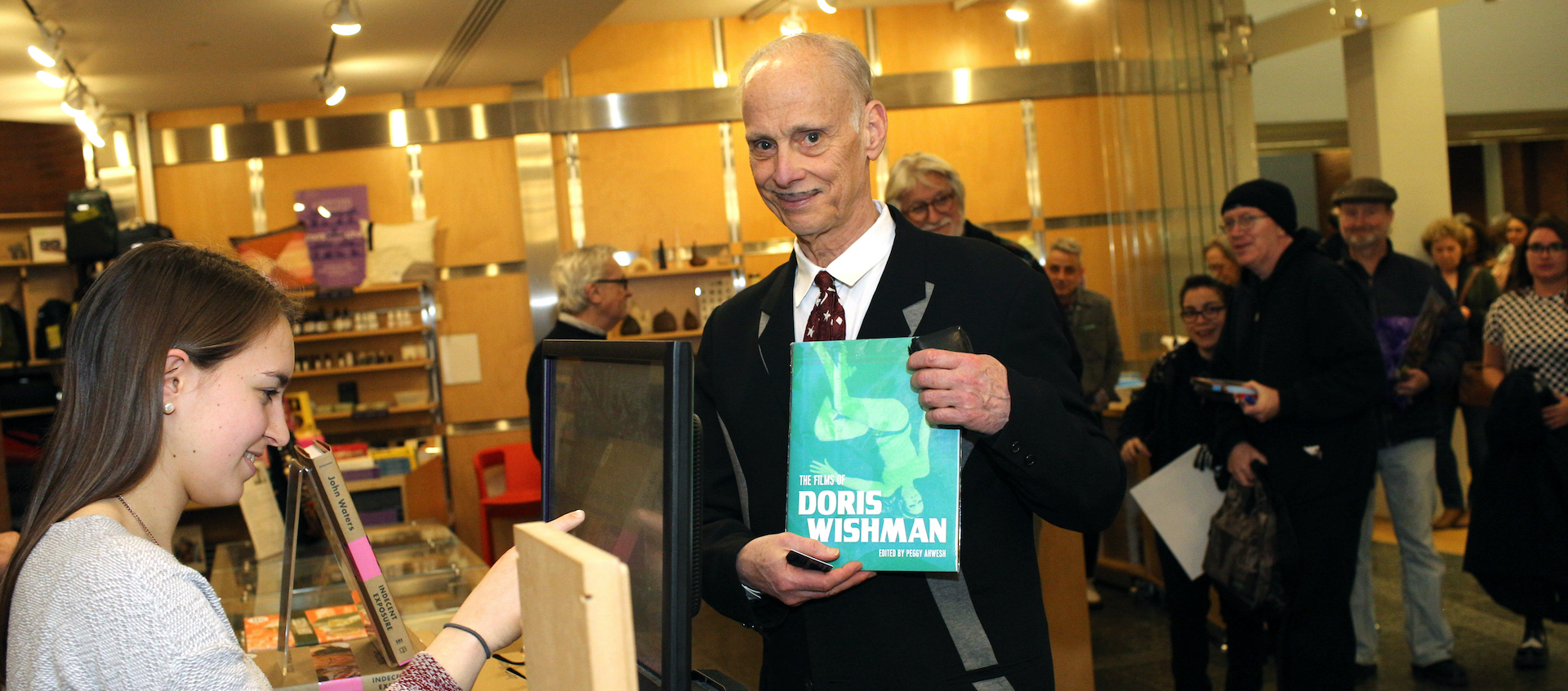 Filmmaker John Waters holds up a copy of a book about Doris Wishman as he purchases it from a young woman clerk in the Wexner Center Store. A line of people stand behind Waters