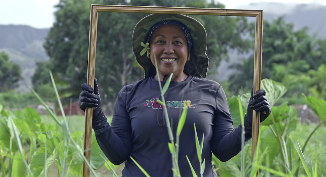 A woman standing in a field of tall grass holds a large gold frame around her face in a scene from a video produced by the Biden-Harris campaign