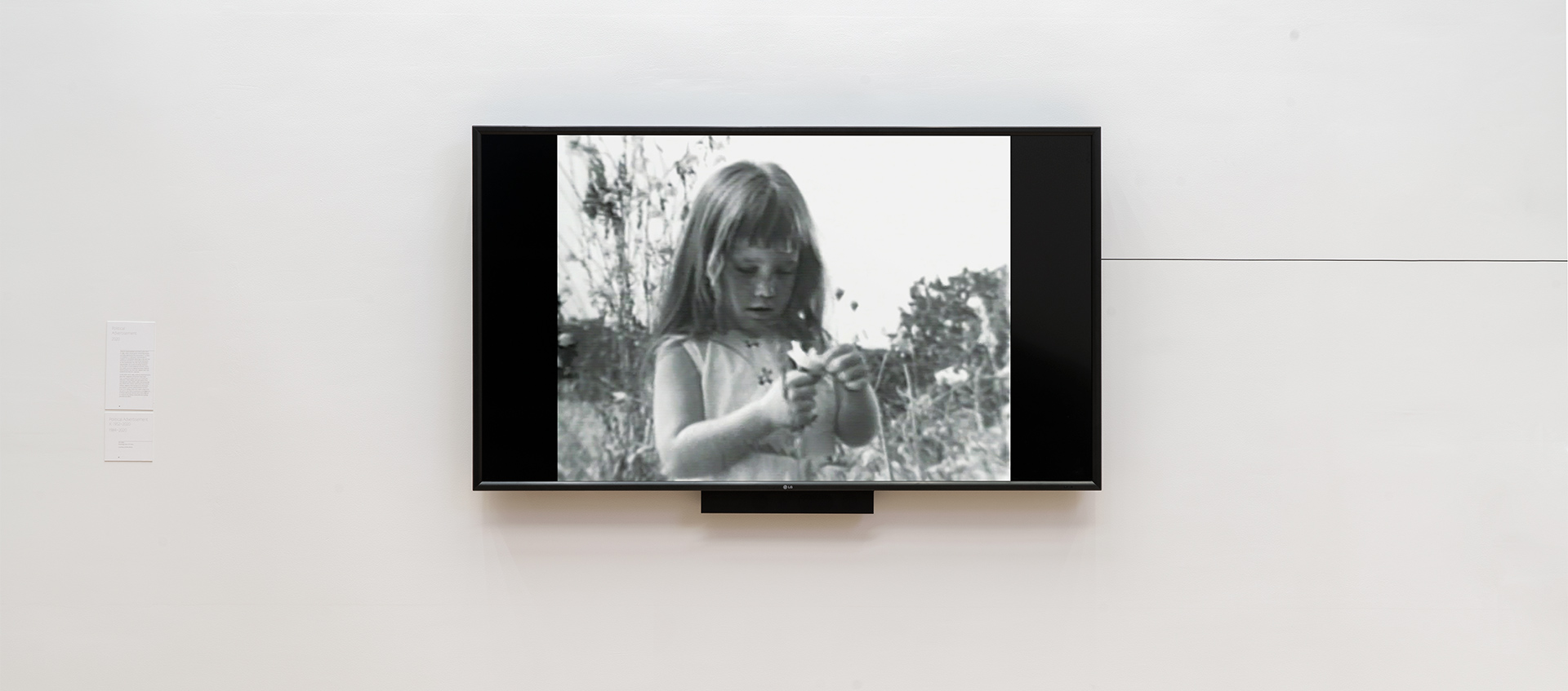A tv mounted to a gallery wall displays a black and white image of a young girl holding a flower.