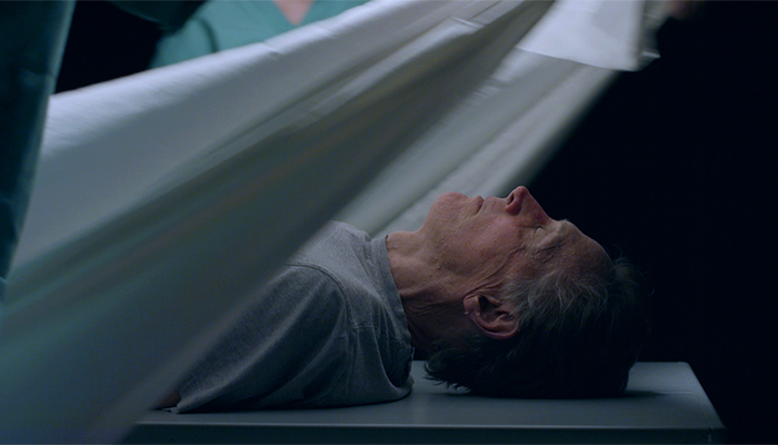 Actor Mark Metcalf, lying down on a flat surface, has a sheet pulled over his head