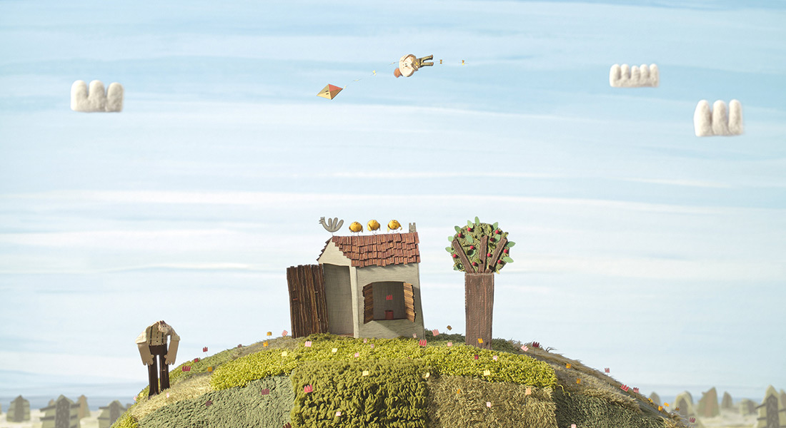 A whimsical landscape crafted from paper, cloth, and fibers depicts a house while a child with their kite flies overhead.