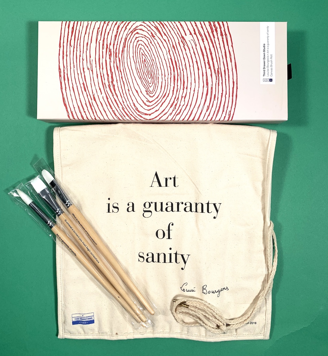 A canvas paintbrush roll bearing a quote from Louise Bourgeois, pictured with the three brushes that come with the roll and the box it all comes in, which bears a spiral design by the artist