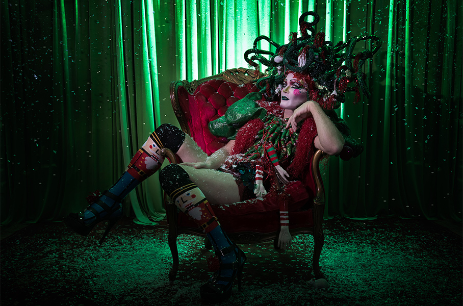 Taylor Mac sits in a red plush chair while wearing festive attire and a snake headdress.
