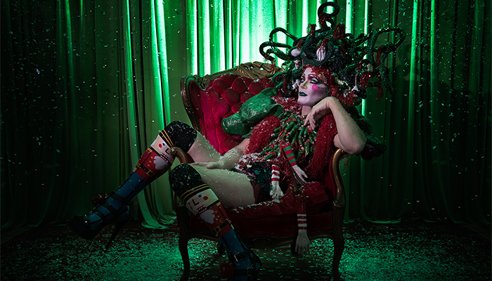 Taylor Mac sits in a red plush chair while wearing festive attire and a snake headdress.
