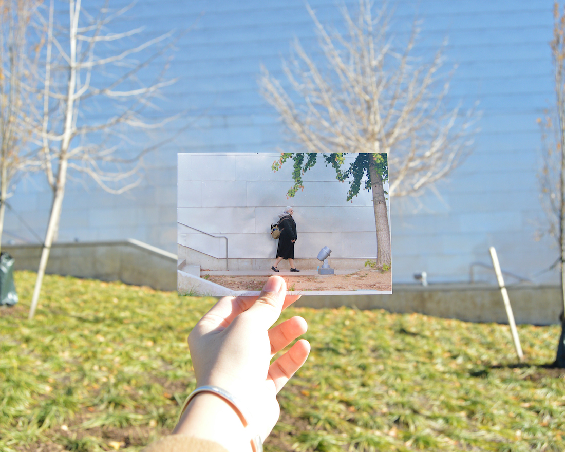 On a grassy hill with a white building in the background, a woman's hand holds up a photo print of a woman in a dark dress and coat walking through the location where the woman holding the photo is standing, so the print almost appears to blend into the surroundings